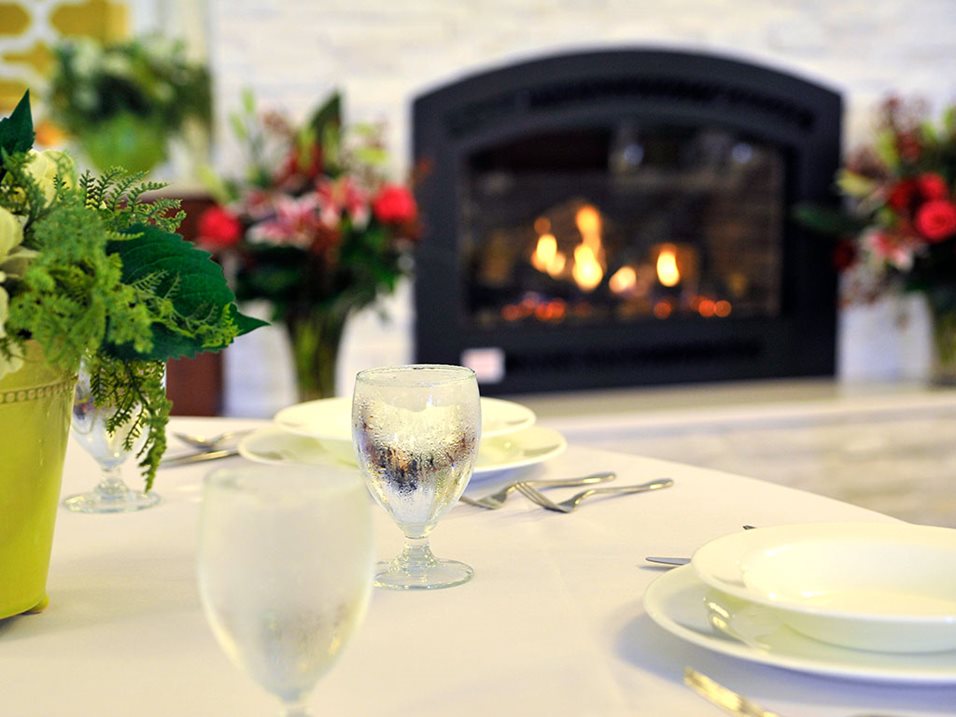 Table set with plates, glasses, and flowers in front of a cozy fireplace.