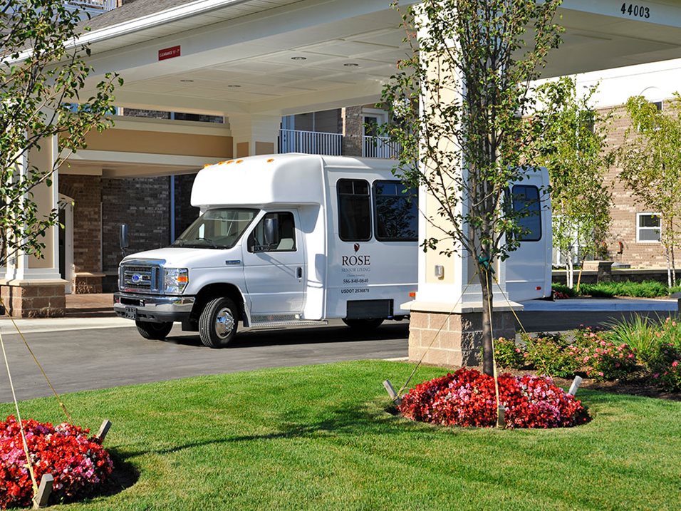 A white shuttle bus parked under the porch of a senior living facility on a sunny day.
