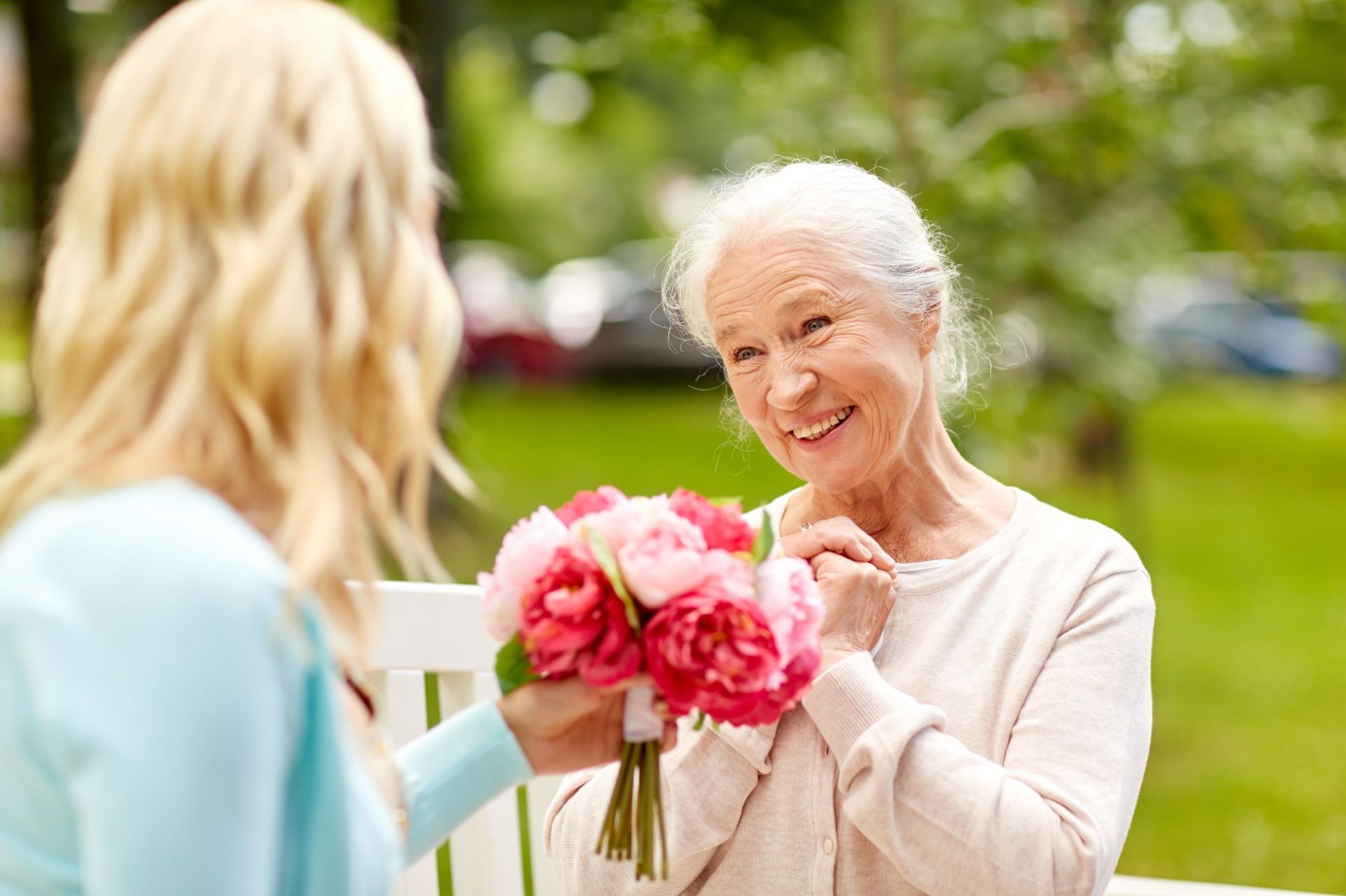 Elderly woman smiling and receiving flowers from younger woman in a park setting.