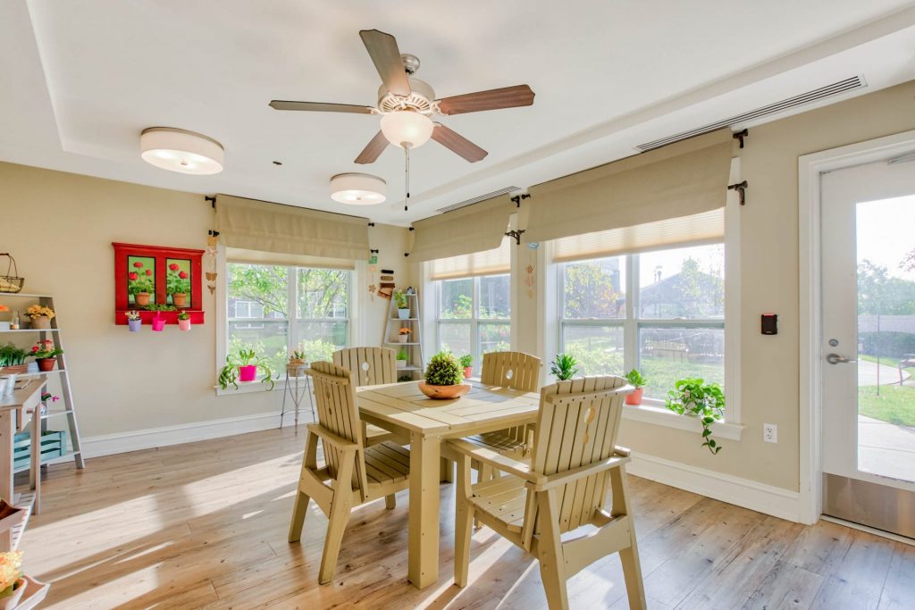 Bright room with wooden table and chairs, ceiling fan, and plants on shelves and windowsills`