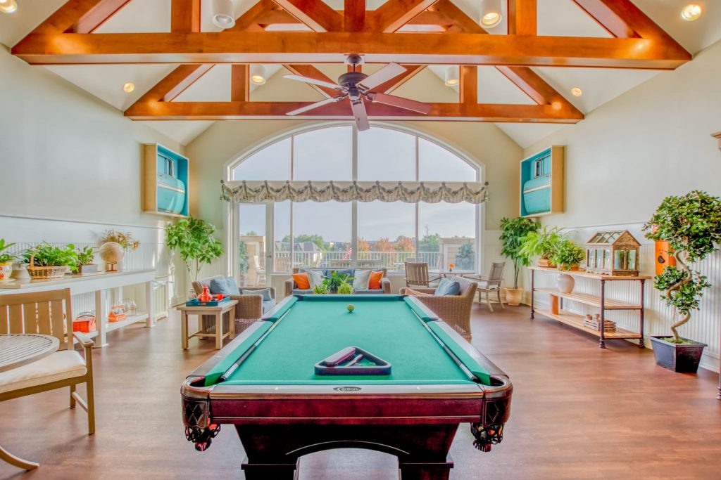 Spacious recreational room with exposed beams, billiard table, plants, bench, sofas, large windows.