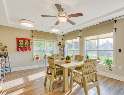 Bright airy kitchen with wooden dining set, ceiling fan, and large windows with plants.